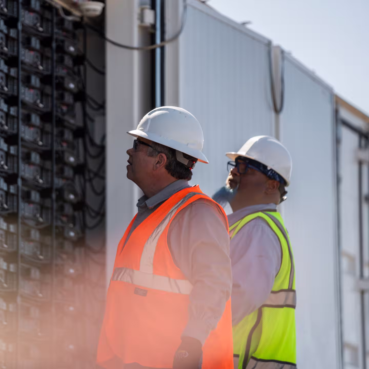 Two workers wearing hard hats and reflective vests inspecting electrical equipment outdoors.