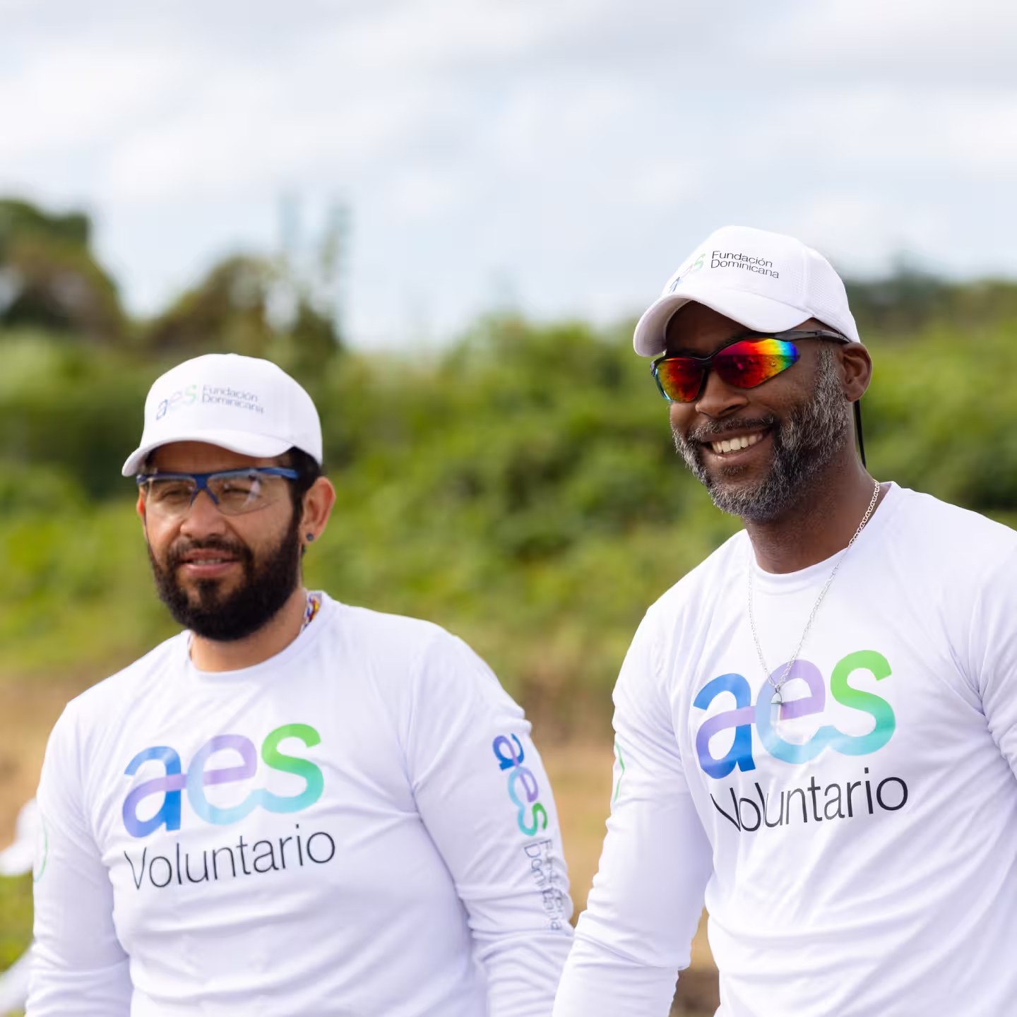 Two men wearing white 'aes Voluntario' shirts and white caps smiling outdoors with a blurred green background.
